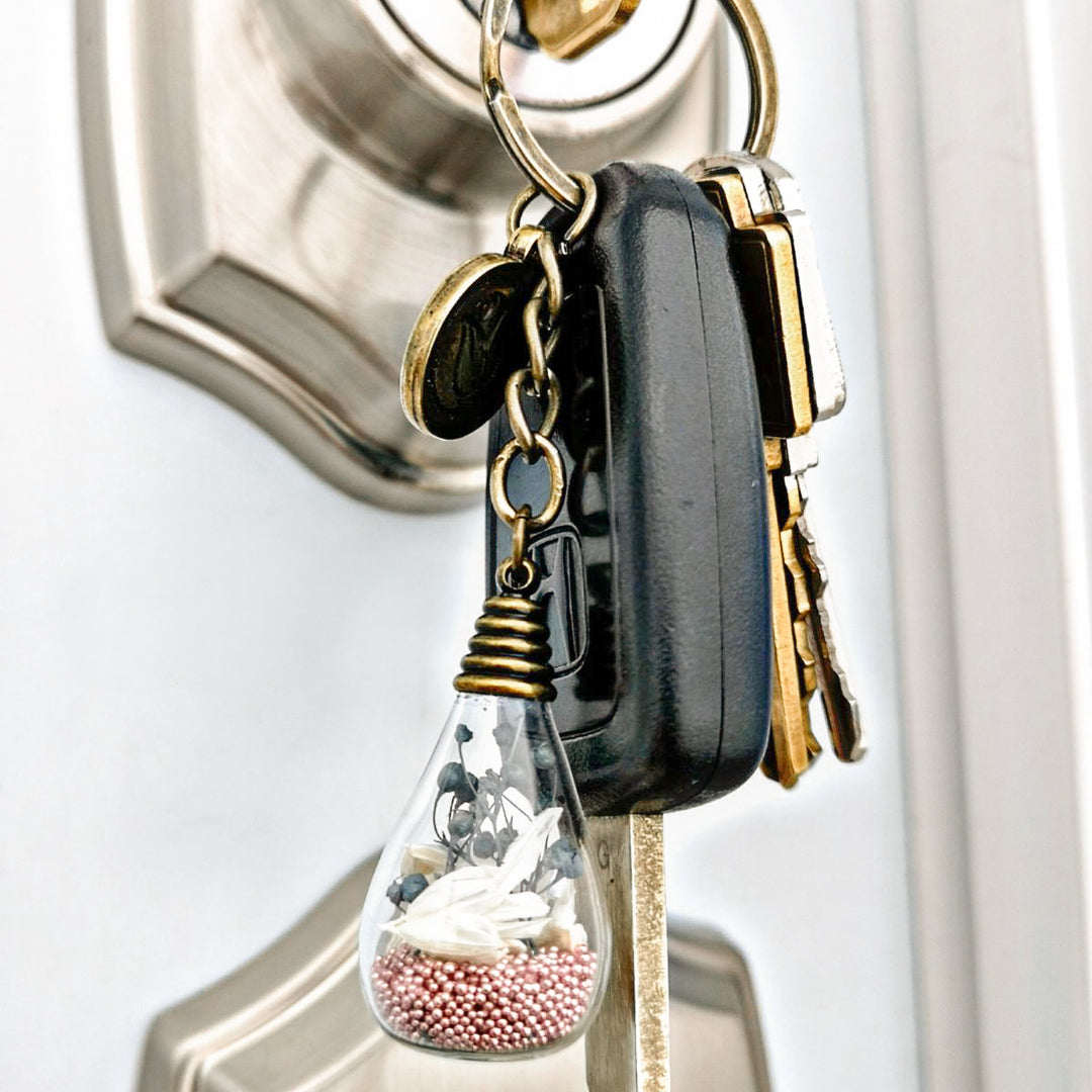 Porte-clé bronze antique avec fleurs, gypsophiles gris et billes rose. Création florale évoquant la paix intérieure et l'équilibre émotionnel.
