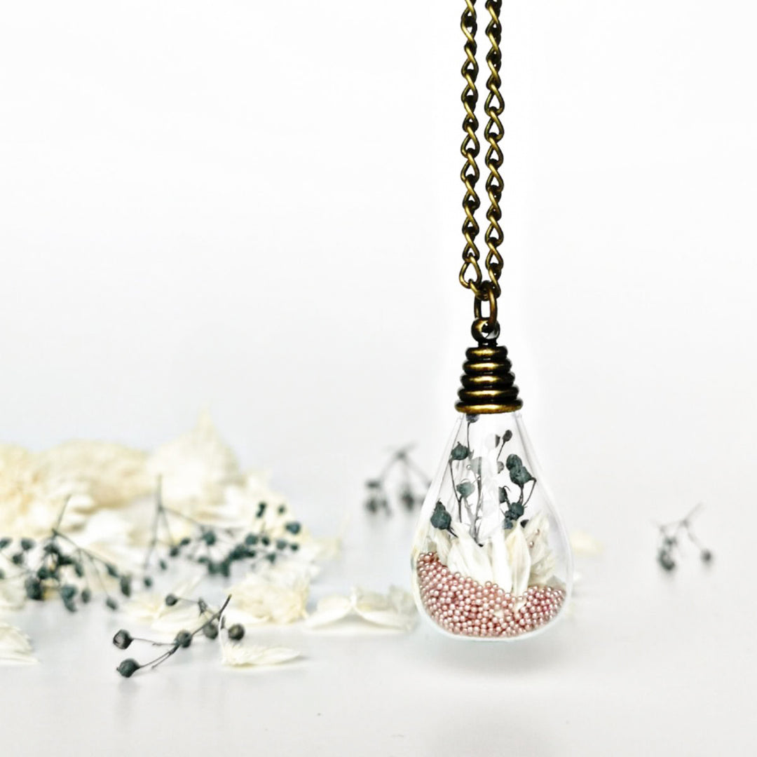 Pendentif fait main avec fleurs crème, gypsophiles gris et billes rose. Création florale évoquant la paix intérieure et l'équilibre émotionnel.
