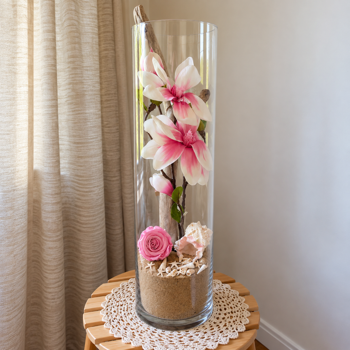 Arrangement floral avec magnolias roses, rose éternelle rose, bois flotté, sable et coquillages