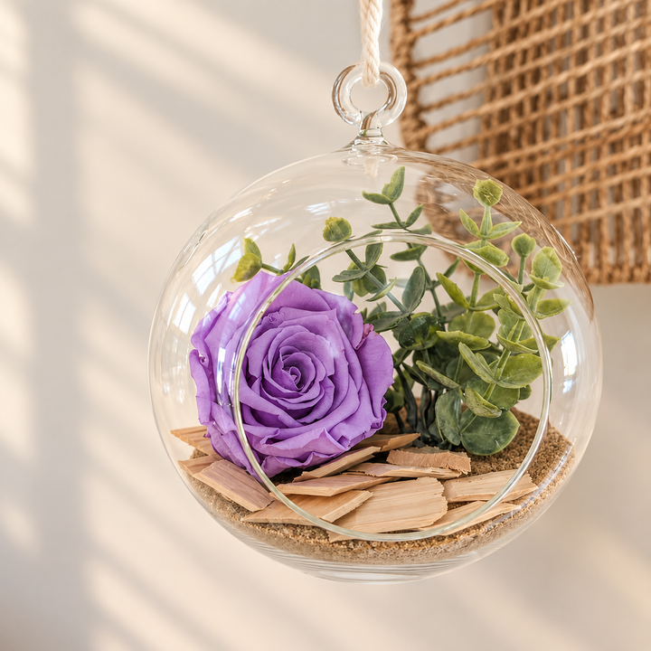 Arrangement floral dans un vase boule suspendu en verre avec rose mauve, eucalyptus et morceaux de bois sur sable.