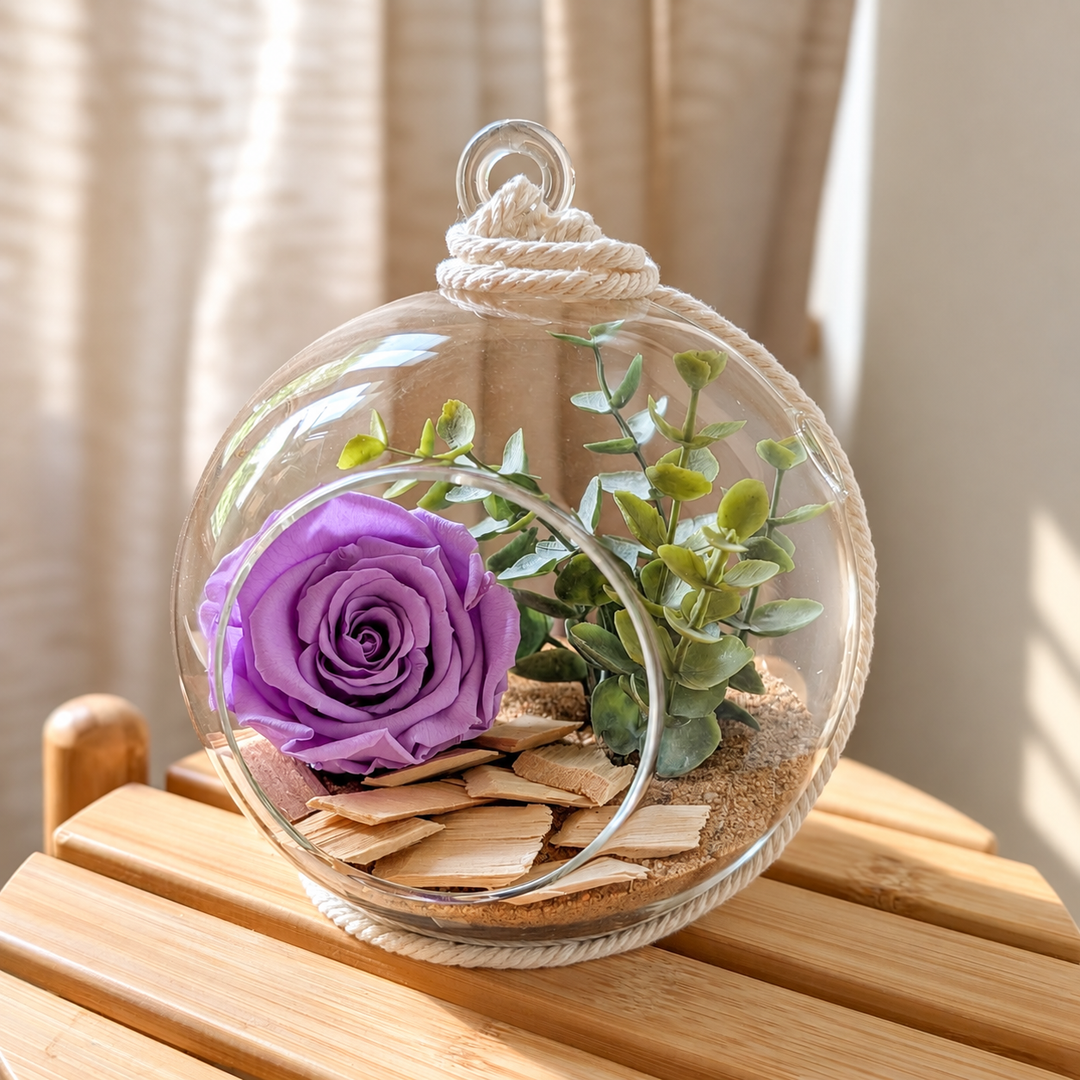 Arrangement floral dans un vase boule en verre avec rose mauve, eucalyptus, corde en macramé et morceaux de bois sur sable