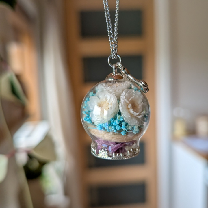 Collier contenant les poils d'un chien décédé, avec fleur de gypsophiles bleues et hélichrysum blanches dans un globe de verre et une breloque en forme d'os