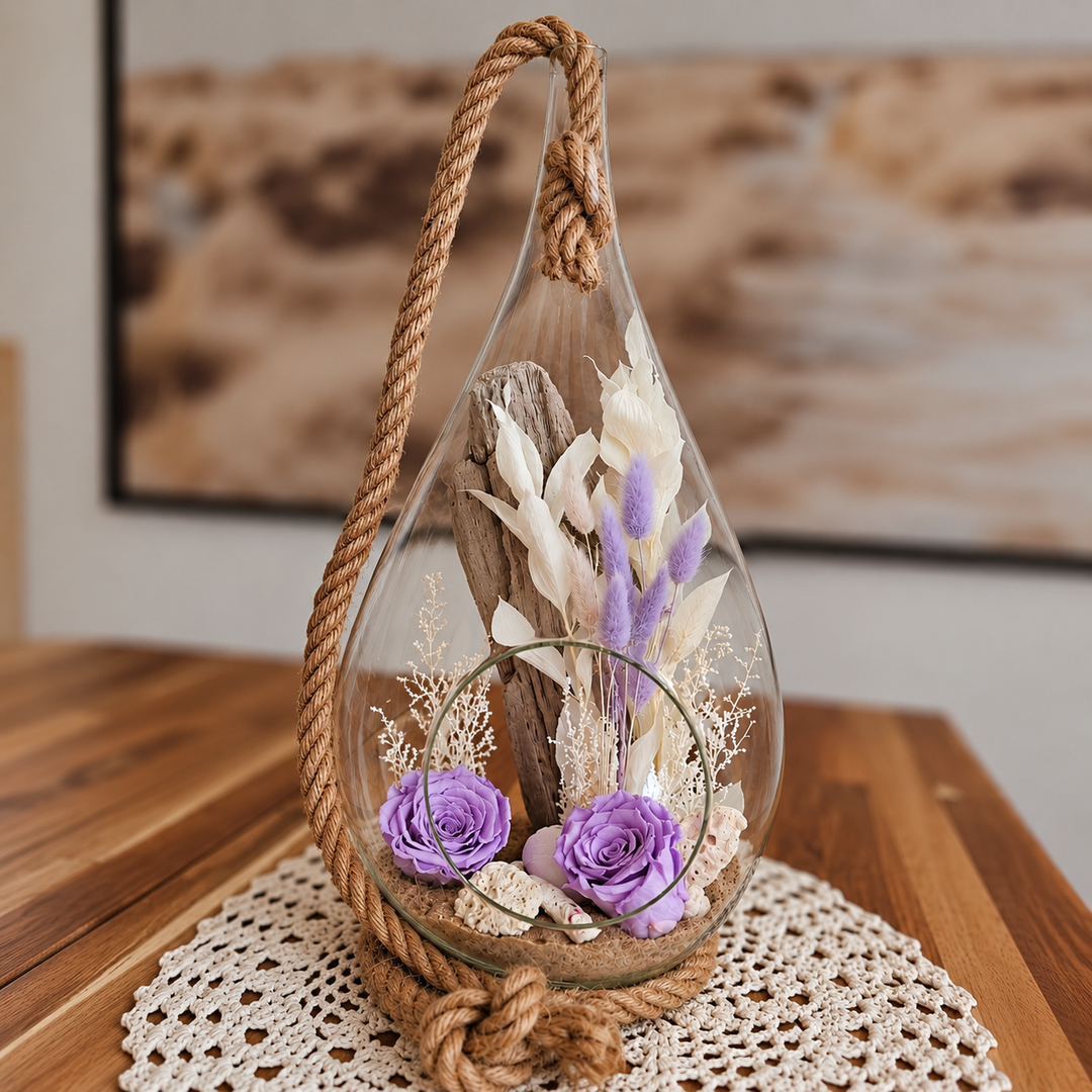 Arrangement floral hommage avec roses éternelles mauves, bois flotté et coquillages dans un vase goutte