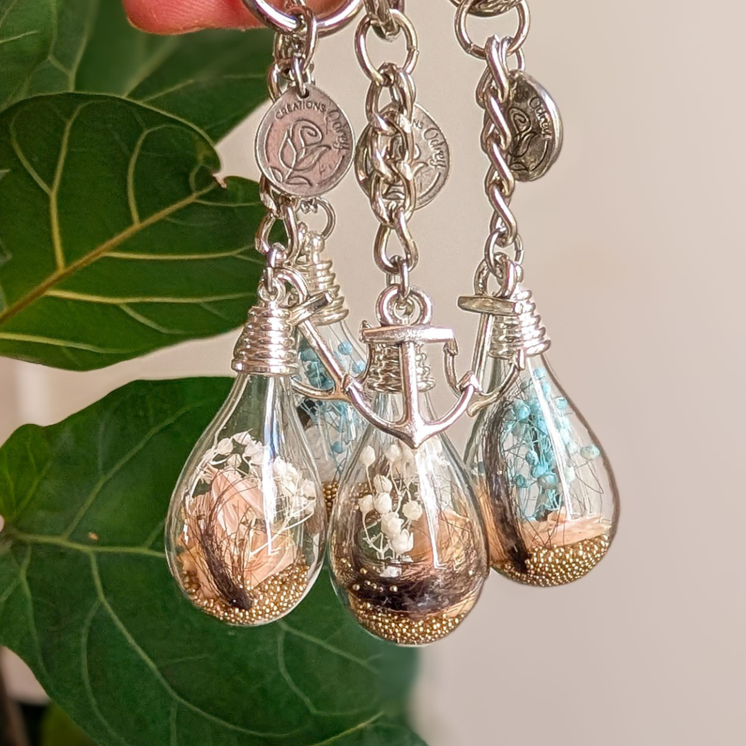 Quatre porte-clés souvenir en goutte de verre avec une mèche de cheveux, des fleurs séchées et une breloque ancre de bateau