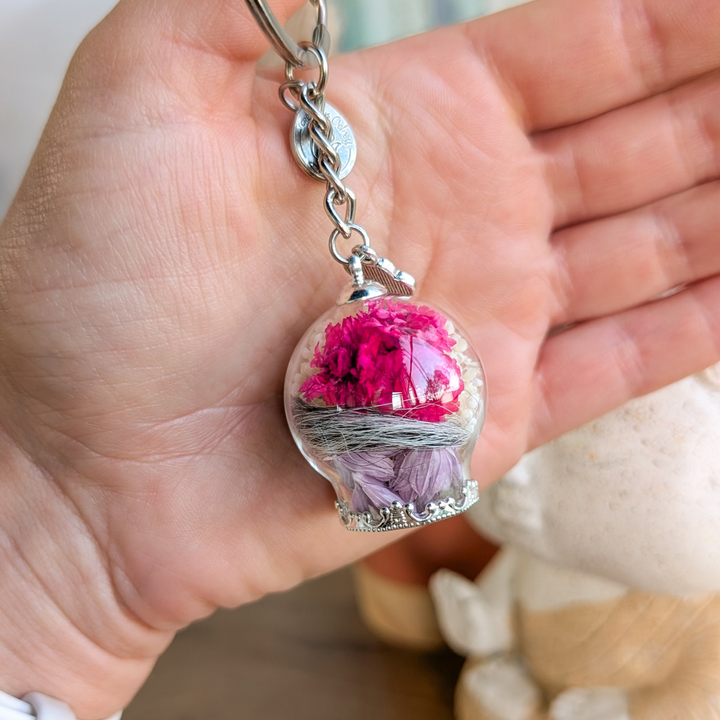 Porte-clé souvenir en acier inoxydable argenté avec poils d’animal, fleurs fuchsia et mauves dans un globe de verre